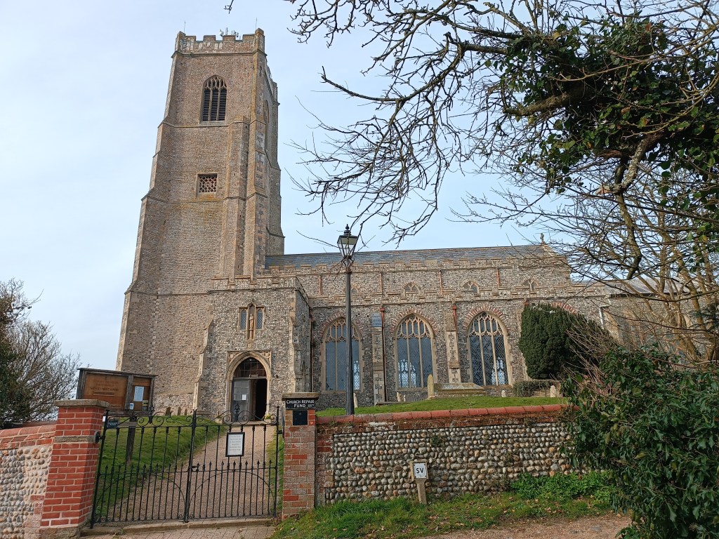 A medieval church with a tall stone tower, surrounded by trees and a stone wall, featuring a pathway leading to the entrance.