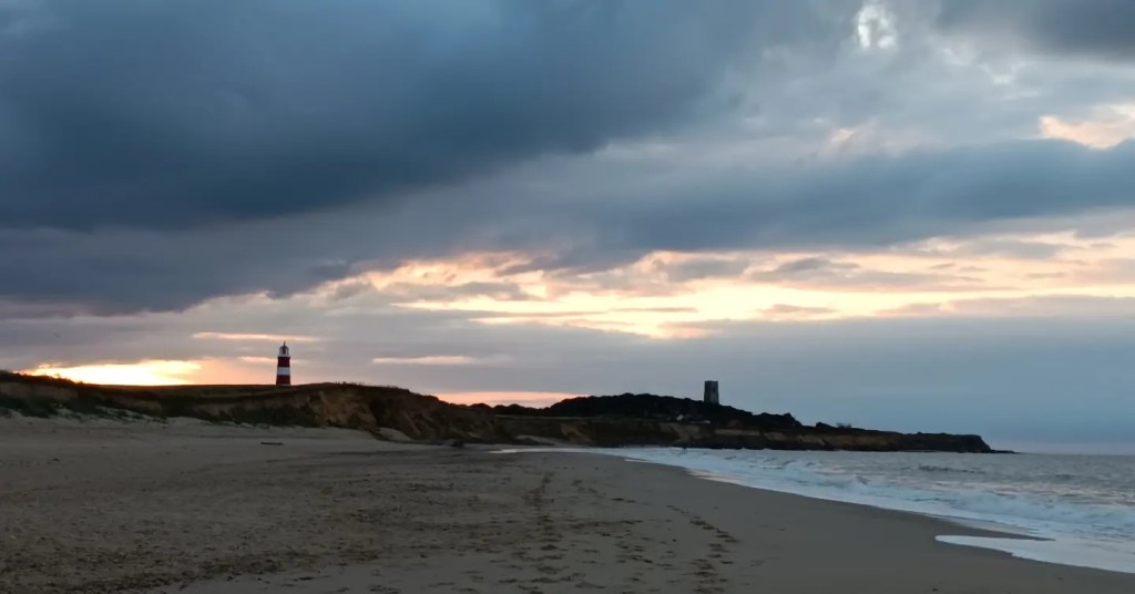 A scenic beach at sunset, featuring a striped lighthouse in the distance and dramatic clouds in the sky.